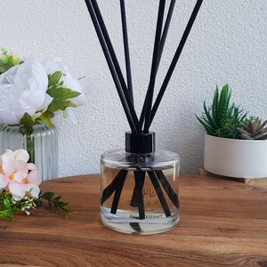 May include: A clear glass bottle diffuser with black reeds and a black label that says "Reed Diffuser". The bottle is sitting on a wooden surface with a white wall in the background.