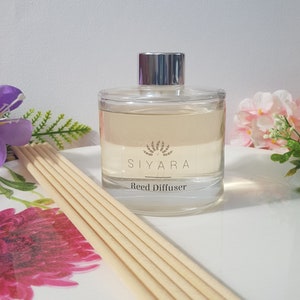 May include: A clear glass bottle with a silver lid filled with a light yellow liquid. The bottle has a label that says "Siyara Reed Diffuser".  The bottle is sitting on a white surface with wooden sticks.