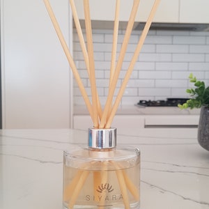 May include: A clear glass bottle with a silver lid and wooden sticks. The bottle contains a light yellow liquid and has a label that reads "Siyara Reed Diffuser".