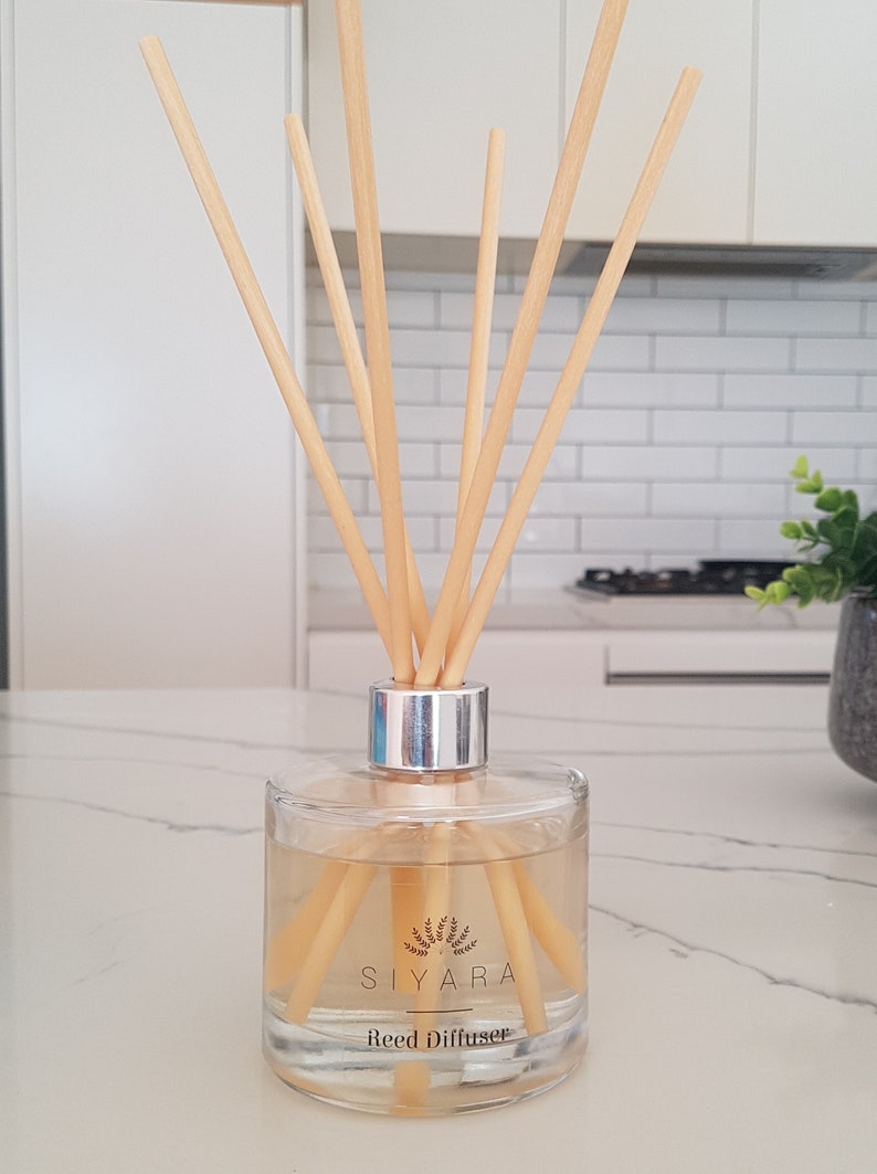 May include: A clear glass bottle with a silver lid and wooden sticks. The bottle contains a light yellow liquid and has a label that reads "Siyara Reed Diffuser".