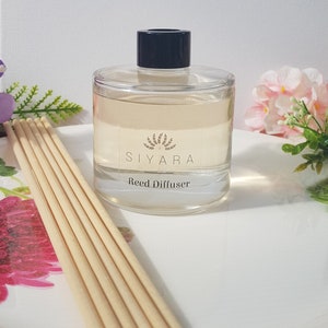 May include: A clear glass bottle with a black lid and a label that reads "Siyara Reed Diffuser". The bottle is filled with a light yellow liquid and is sitting on a white surface. There are wooden sticks in the bottle.
