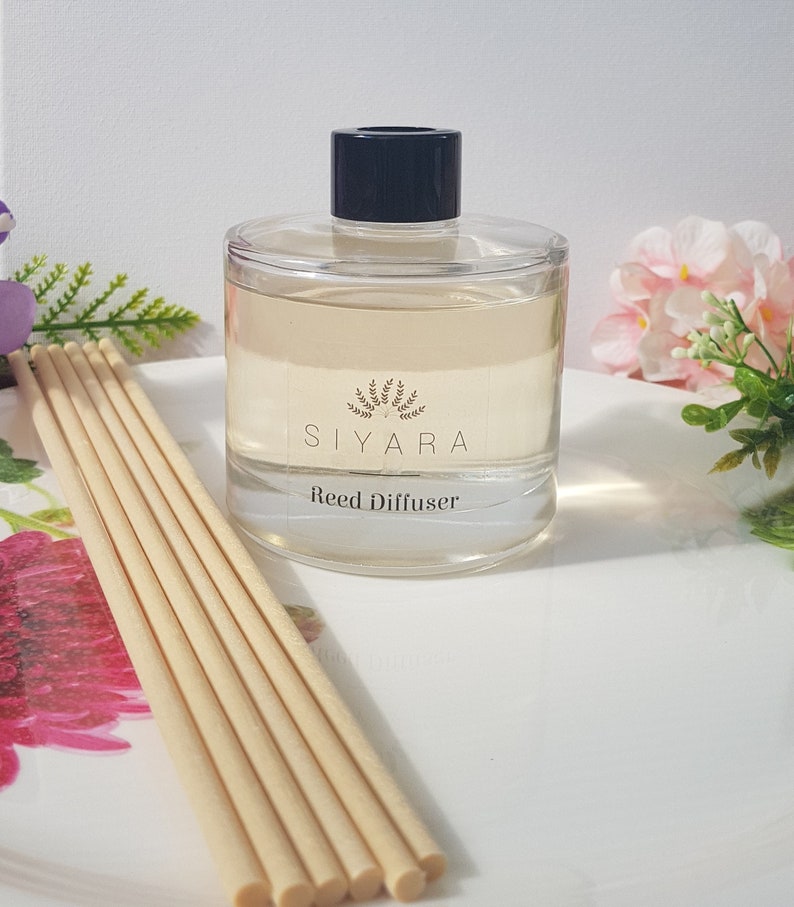 May include: A clear glass bottle with a black lid and a label that reads "Siyara Reed Diffuser". The bottle is filled with a light yellow liquid and is sitting on a white surface. There are wooden sticks in the bottle.