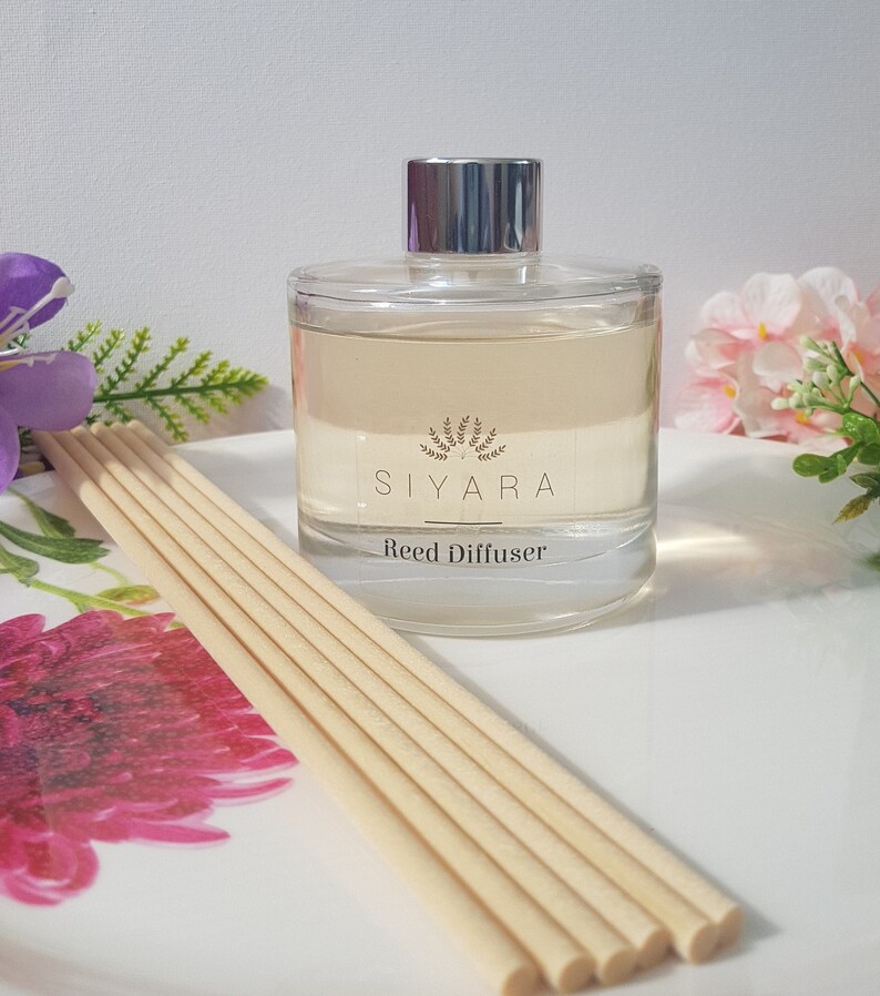 May include: A clear glass bottle with a silver lid filled with a light yellow liquid. The bottle has a label that says "Siyara Reed Diffuser".  The bottle is sitting on a white surface with wooden sticks.