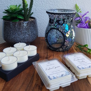 May include: A black mosaic wax warmer with a teal blue interior, surrounded by six white tea light candles in a black holder, and two wax melts in clear plastic containers. The wax melts are labelled "Lychee Guava" and "Raspberry & Vanilla".