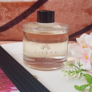 May include: A clear glass bottle with a black lid and a white label that reads "Siyara Reed Diffuser". The bottle is sitting on a white surface with black reed sticks and a pink flower.