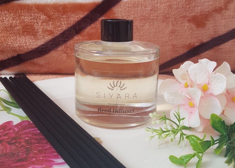 May include: A clear glass bottle with a black lid and a white label that reads "Siyara Reed Diffuser". The bottle is sitting on a white surface with black reed sticks and a pink flower.