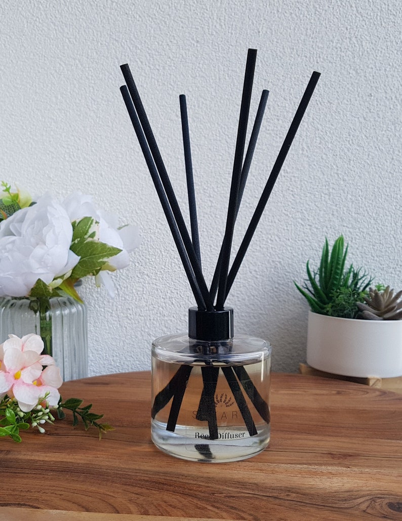May include: A clear glass bottle diffuser with black reeds and a black label that says "Reed Diffuser". The bottle is sitting on a wooden surface with a white wall in the background.