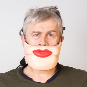 Op de afbeelding: Een man draagt ​​een wit gezichtsmasker met een rode mond en een zwarte band. Het masker bedekt zijn mond en kin.