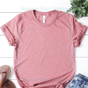 May include: A dusty rose coloured t-shirt with rolled up sleeves, a pair of light blue jeans, and brown sandals on a white wooden background.