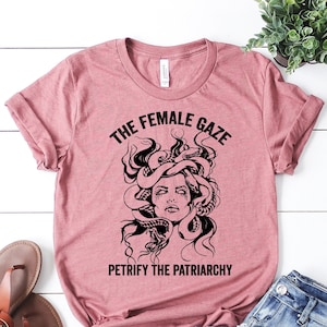 May include: A pink t-shirt with a black graphic of a woman with snakes for hair. The text on the shirt reads "The Female Gaze" and "Petrify the Patriarchy".