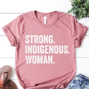 May include: A pink t-shirt with the text "STRONG. INDIGENOUS. WOMAN." printed in white on the front. The shirt is laid out on a white wooden surface with a pair of brown sandals and blue jeans.