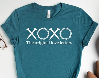 May include: A teal blue t-shirt with a white graphic that says "XOXO The original love letters". The shirt is laid flat on a white surface with other items around it, including a pair of glasses, a notebook, a pen, a small jewelry dish, a scarf, and a pair of brown shoes.