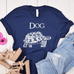 May include: A navy blue t-shirt with a white graphic of a turtle and the word "DOG" above it. The shirt is laid out on a white wooden surface with a pair of light brown heeled sandals and light blue jeans.