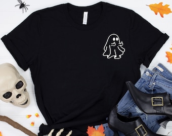 May include: Black t-shirt with a white ghost graphic, styled with blue jeans, black witch shoes, and Halloween decorations including a skull, broom, and spiders. Autumn leaves and candy corn complete the festive scene.
