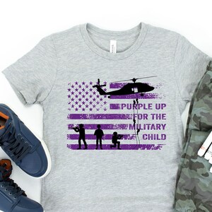 K&ouml;nnte beinhalten: Ein graues T-Shirt mit einem "Purple Up for the Military Child"-Design. Das Design zeigt einen Hubschrauber, Soldaten und eine amerikanische Flagge.
