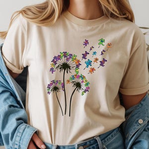May include: Beige t-shirt featuring a colorful dandelion design. The dandelion heads are composed of purple, green, and blue flowers, with small, colorful figures blowing away from the flower heads. The shirt has short sleeves and a crew neck.