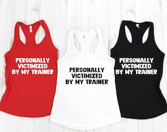 Könnte beinhalten: Drei Tanktops mit dem Aufdruck "PERSONALLY VICTIMIZED BY MY TRAINER". Die Tanktops sind rot, weiß und schwarz.