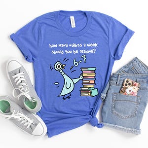 K&ouml;nnte beinhalten: Blaues T-Shirt mit einem Cartoon-Tauben-Motiv, das B&uuml;cher h&auml;lt, und dem Text "Wie viele N&auml;chte pro Woche solltest du lesen?" mit der Antwort "6-7". Graue Turnschuhe und Jeansshorts sind ebenfalls zu sehen.