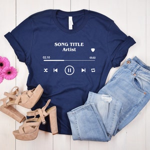 May include: A navy blue t-shirt with a white graphic of a music player with the text "SONG TITLE Artist" and the time 02:10 to 03:02. The t-shirt is laying on a white wooden surface with a pair of light brown heeled sandals and light blue jeans.