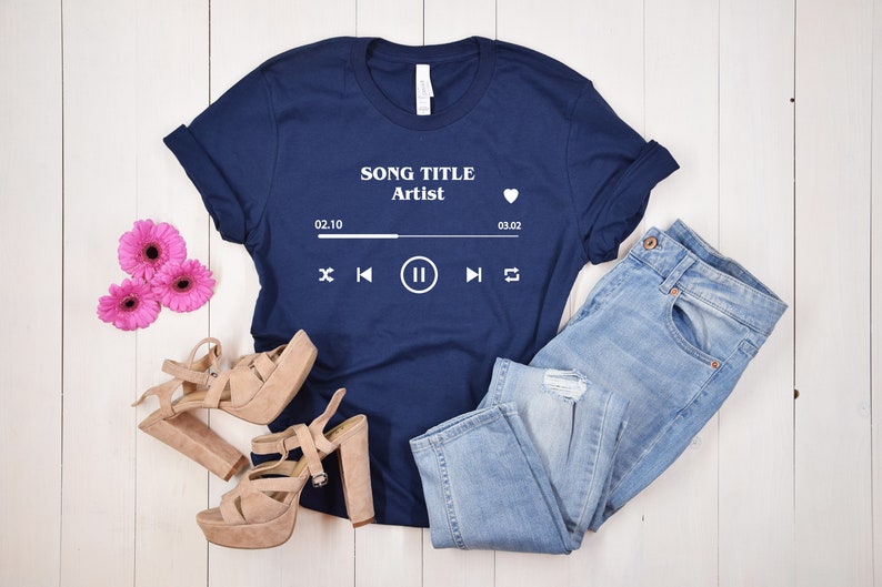 May include: A navy blue t-shirt with a white graphic of a music player with the text "SONG TITLE Artist" and the time 02:10 to 03:02. The t-shirt is laying on a white wooden surface with a pair of light brown heeled sandals and light blue jeans.
