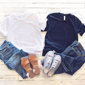 May include: Two blank t-shirts, one white and one black, are laid out on a whitewashed wood background.  A pair of blue jeans and a pair of brown sandals are placed next to the white t-shirt.  A pair of blue jeans and a pair of grey trainers are placed next to the black t-shirt.
