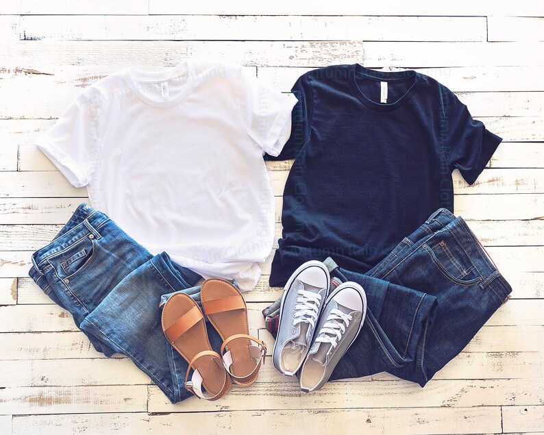 May include: Two blank t-shirts, one white and one black, are laid out on a whitewashed wood background.  A pair of blue jeans and a pair of brown sandals are placed next to the white t-shirt.  A pair of blue jeans and a pair of grey trainers are placed next to the black t-shirt.