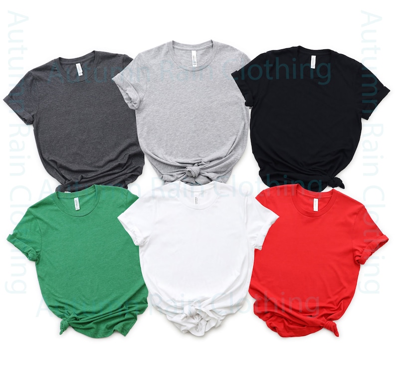 May include: Six blank t-shirts in different colours: dark grey, light grey, black, green, white, and red. The shirts are folded and laid out on a white background.