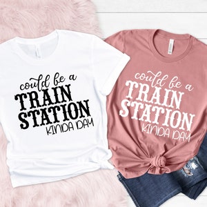May include: Two t-shirts with the text "could be a train station kinda day" printed on them. One t-shirt is white with black text and the other is pink with white text.