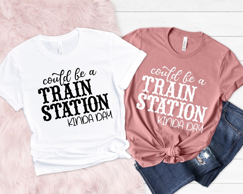 May include: Two t-shirts with the text "could be a train station kinda day" printed on them. One t-shirt is white with black text and the other is pink with white text.