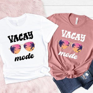 May include: Two t-shirts, one white and one dusky pink, each with the words "VACAY mode" above a graphic of sunglasses with a sunset and palm tree design. The white shirt is knotted at the waist, and the other is paired with blue jeans.
