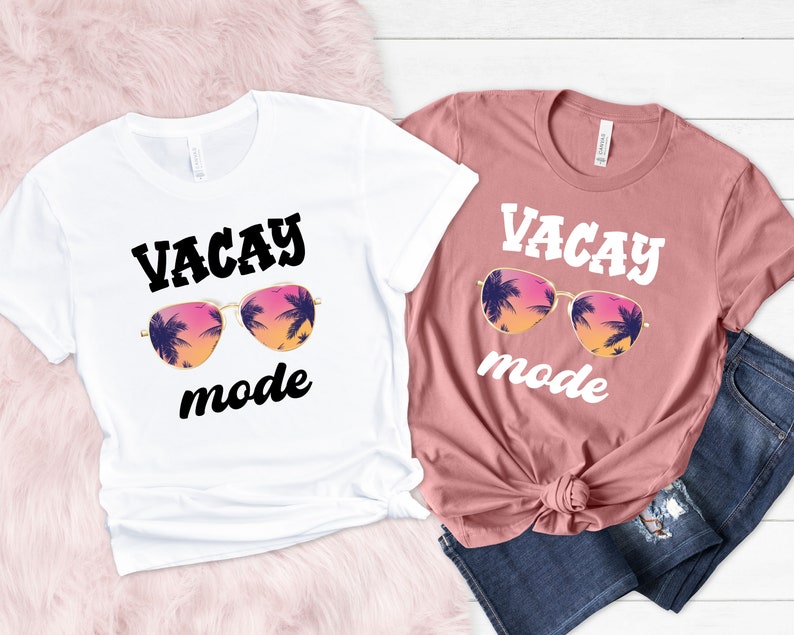 May include: Two t-shirts, one white and one dusky pink, each with the words "VACAY mode" above a graphic of sunglasses with a sunset and palm tree design. The white shirt is knotted at the waist, and the other is paired with blue jeans.