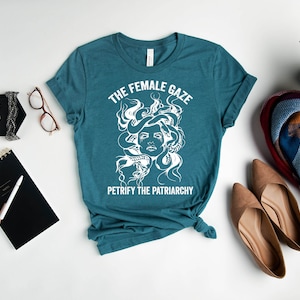 May include: Teal blue t-shirt with a white graphic of Medusa's head with snakes for hair. The text "The Female Gaze" and "Petrify the Patriarchy" is printed on the shirt.