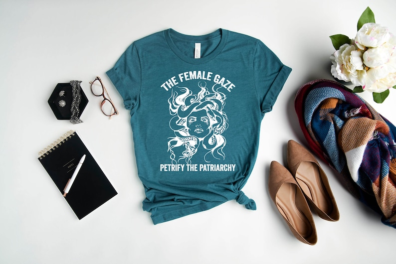 May include: Teal blue t-shirt with a white graphic of Medusa's head with snakes for hair. The text "The Female Gaze" and "Petrify the Patriarchy" is printed on the shirt.