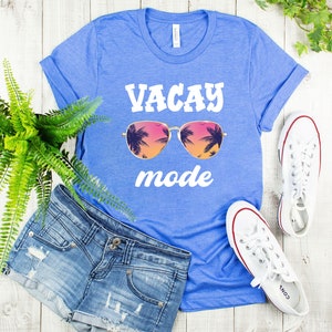 May include: A blue t-shirt with the words "VACAY mode" in white, featuring sunglasses with a tropical sunset design. The shirt is paired with denim shorts and white trainers, suggesting a holiday theme.