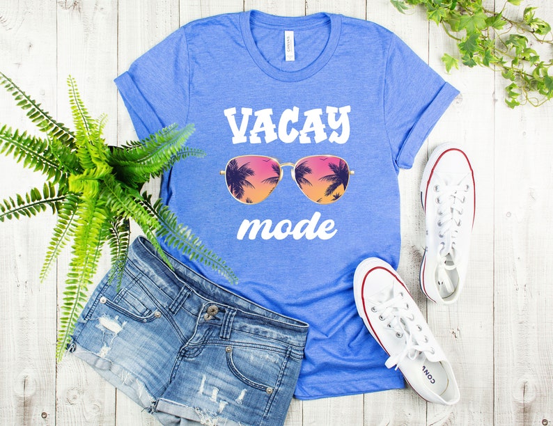May include: A blue t-shirt with the words "VACAY mode" in white, featuring sunglasses with a tropical sunset design. The shirt is paired with denim shorts and white trainers, suggesting a holiday theme.