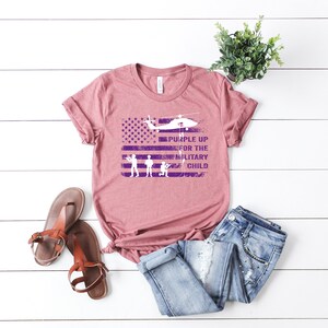 K&ouml;nnte beinhalten: Ein mauvefarbenes T-Shirt mit einem abgenutzten, violetten amerikanischen Flaggenmuster. Die Flagge hat einen dar&uuml;ber fliegenden Hubschrauber und den Text "Purple Up for the Military Child".