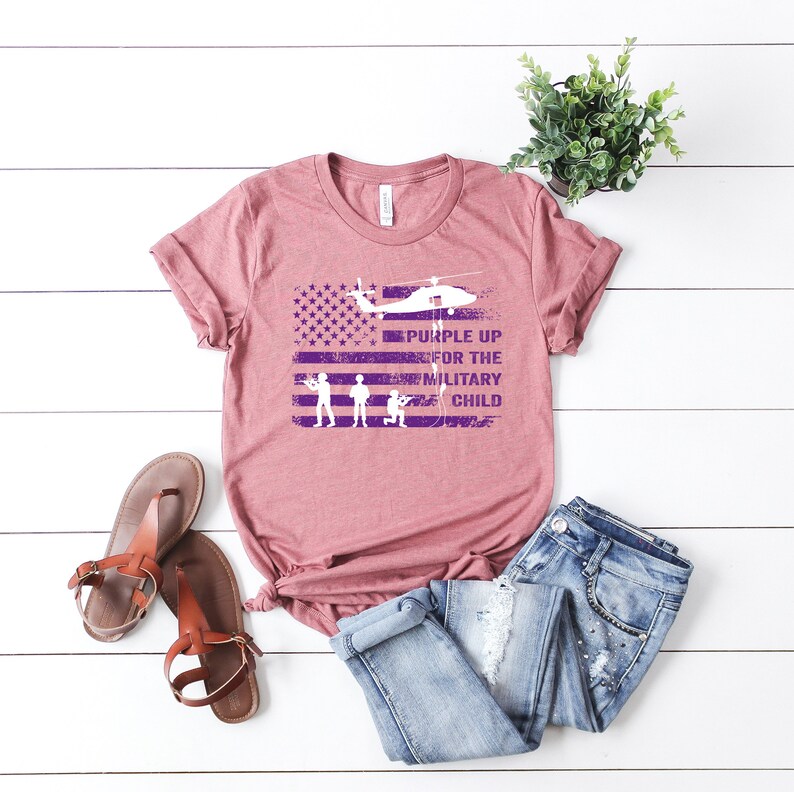K&ouml;nnte beinhalten: Ein mauvefarbenes T-Shirt mit einem abgenutzten, violetten amerikanischen Flaggenmuster. Die Flagge hat einen dar&uuml;ber fliegenden Hubschrauber und den Text "Purple Up for the Military Child".