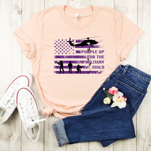 K&ouml;nnte beinhalten: Ein pfirsichfarbenes T-Shirt mit einem abgenutzten amerikanischen Flaggen-Design und dem Text "Purple Up for the Military Child" in Lila. Die Flagge hat einen Hubschrauber, der dar&uuml;ber fliegt, und drei Soldaten in Silhouette.