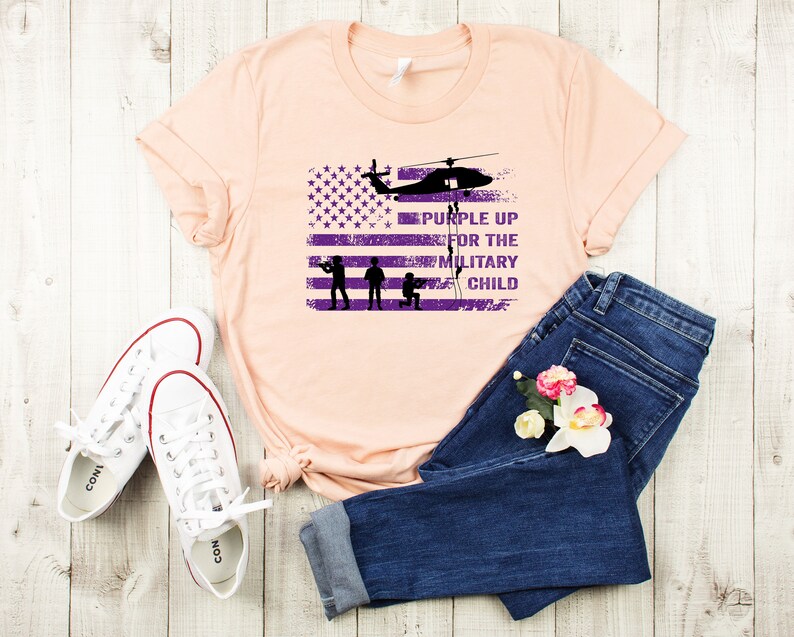 K&ouml;nnte beinhalten: Ein pfirsichfarbenes T-Shirt mit einem abgenutzten amerikanischen Flaggen-Design und dem Text "Purple Up for the Military Child" in Lila. Die Flagge hat einen Hubschrauber, der dar&uuml;ber fliegt, und drei Soldaten in Silhouette.