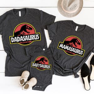 May include: Dark gray t-shirts and a onesie with dinosaur-themed graphics. The shirts say "Dadasaurus" and "Mamasaurus", and the onesie says "Babysaurus". The graphics are in a red and yellow circle.