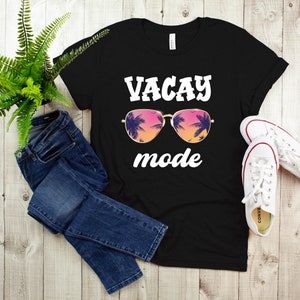 May include: Black t-shirt with the words "VACAY mode" in white, featuring sunglasses with a tropical sunset design. The image also includes blue jeans and white trainers.
