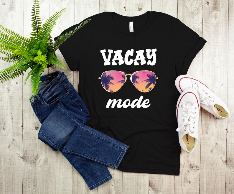 May include: Black t-shirt with the words "VACAY mode" in white, featuring sunglasses with a tropical sunset design. The image also includes blue jeans and white trainers.