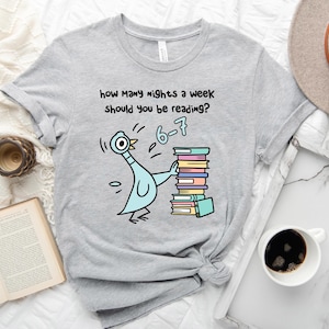 K&ouml;nnte beinhalten: Hellgraues T-Shirt mit einem Cartoon-Pigeon, der B&uuml;cher h&auml;lt. Der Text auf dem Shirt lautet: &bdquo;Wie viele N&auml;chte pro Woche solltest du lesen?&ldquo; mit der Antwort &bdquo;6-7&ldquo;. Eine Kaffeetasse, ein offenes Buch und eine Uhr sind ebenfalls im Bild.