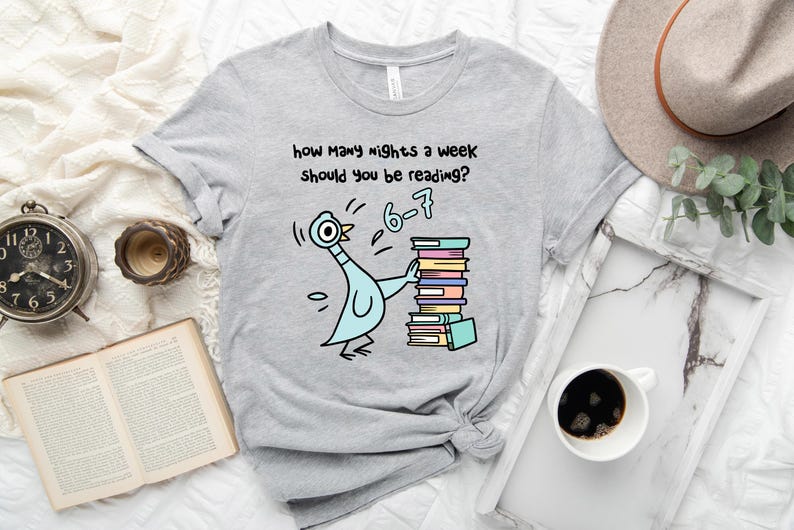 K&ouml;nnte beinhalten: Hellgraues T-Shirt mit einem Cartoon-Pigeon, der B&uuml;cher h&auml;lt. Der Text auf dem Shirt lautet: &bdquo;Wie viele N&auml;chte pro Woche solltest du lesen?&ldquo; mit der Antwort &bdquo;6-7&ldquo;. Eine Kaffeetasse, ein offenes Buch und eine Uhr sind ebenfalls im Bild.