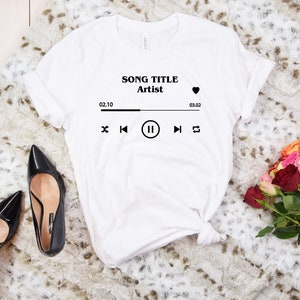 May include: White t-shirt with a black graphic of a music player with the text "SONG TITLE Artist" and a heart icon. The music player graphic includes a play/pause button, a skip forward button, a skip back button, and a repeat button. The time display shows 02.10 and 03.02.