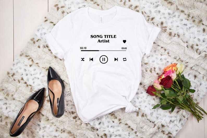 May include: White t-shirt with a black graphic of a music player with the text "SONG TITLE Artist" and a heart icon. The music player graphic includes a play/pause button, a skip forward button, a skip back button, and a repeat button. The time display shows 02.10 and 03.02.