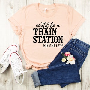 May include: A peach-colored t-shirt with the text "could be a TRAIN STATION kinda day" printed in black. The shirt is laid out on a white wooden surface with a pair of white sneakers and blue jeans.