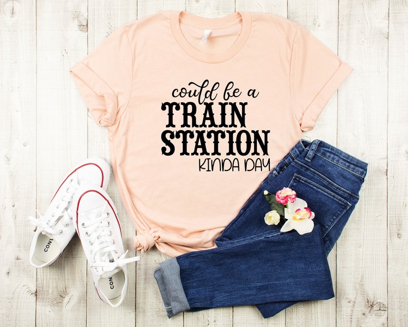 May include: A peach-colored t-shirt with the text "could be a TRAIN STATION kinda day" printed in black. The shirt is laid out on a white wooden surface with a pair of white sneakers and blue jeans.