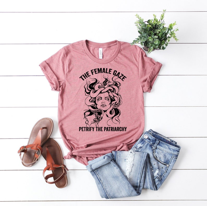 May include: A pink t-shirt with a black graphic of a woman with snakes for hair. The text on the shirt reads "The Female Gaze" and "Petrify the Patriarchy".