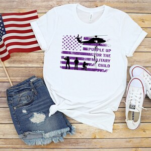 K&ouml;nnte beinhalten: Wei&szlig;es T-Shirt mit einem violetten amerikanischen Flaggen-Design und dem Text "Purple Up for the Military Child".